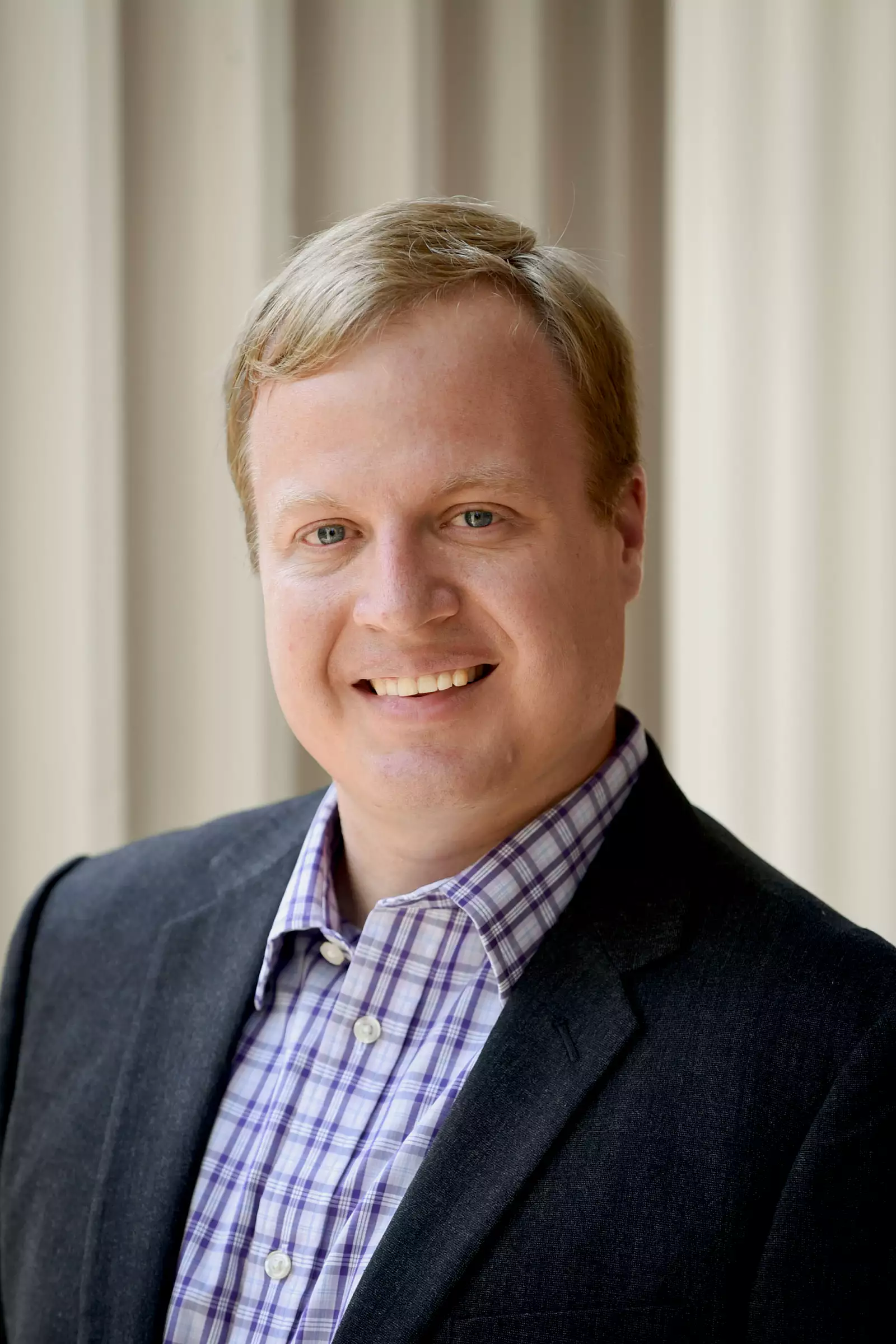 A person with short blond hair is smiling, wearing a dark jacket over a plaid shirt. The background is softly blurred, featuring vertical lines.