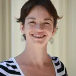 A person with short, dark hair smiles at the camera. They are wearing dangling earrings and a black and white striped top. The background is a solid, light color, creating a simple and clean setting.