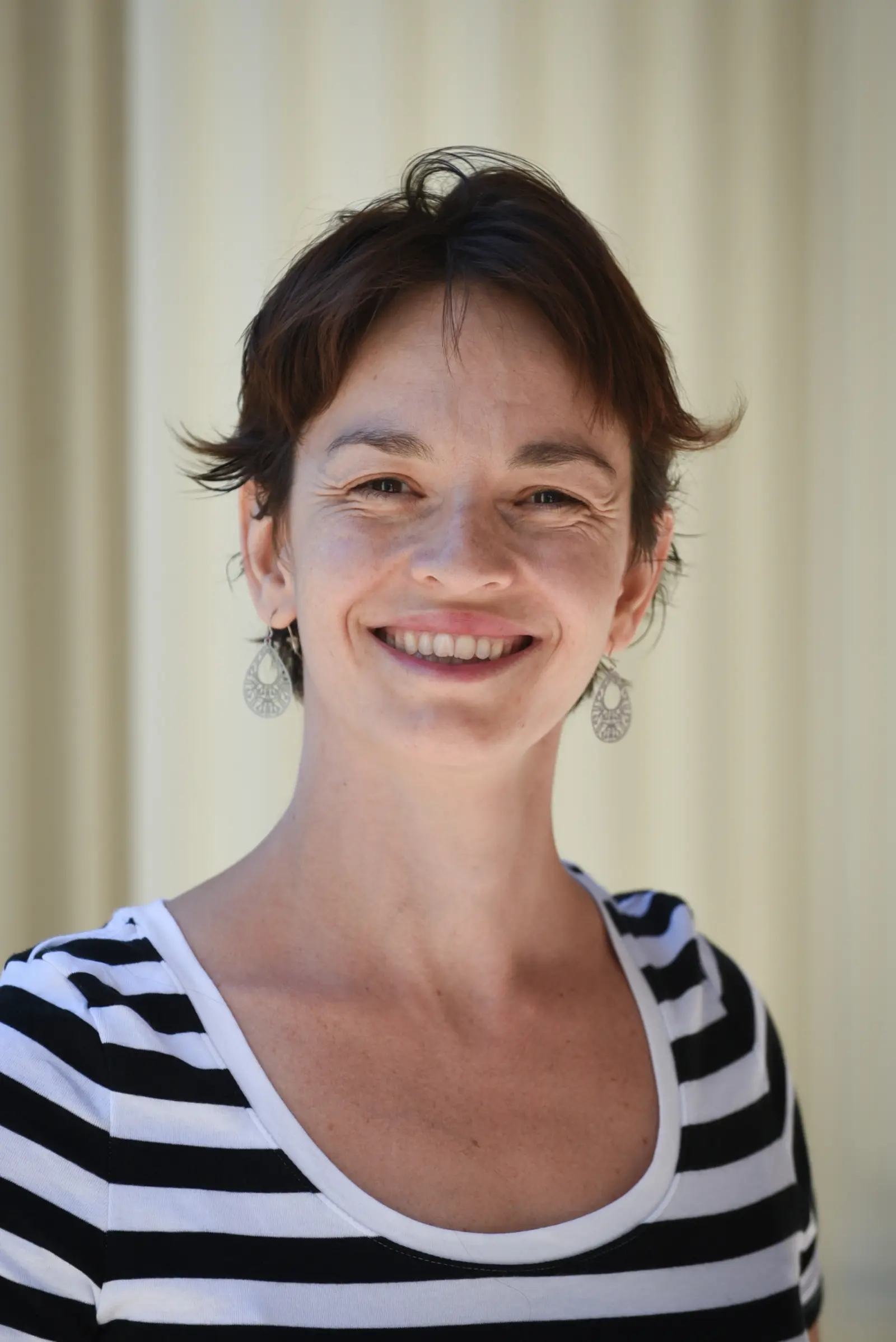A person with short, dark hair smiles at the camera. They are wearing dangling earrings and a black and white striped top. The background is a solid, light color, creating a simple and clean setting.