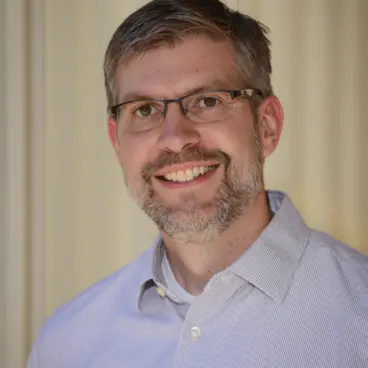 A man with short hair and glasses smiles at the camera, wearing a light striped button-up shirt. The background is softly blurred, creating a neutral and warm tone.