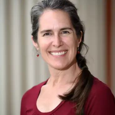A woman with long dark hair, partially gray, smiles at the camera. She is wearing a long-sleeve maroon shirt and small earrings. The background is blurred with soft vertical lines.