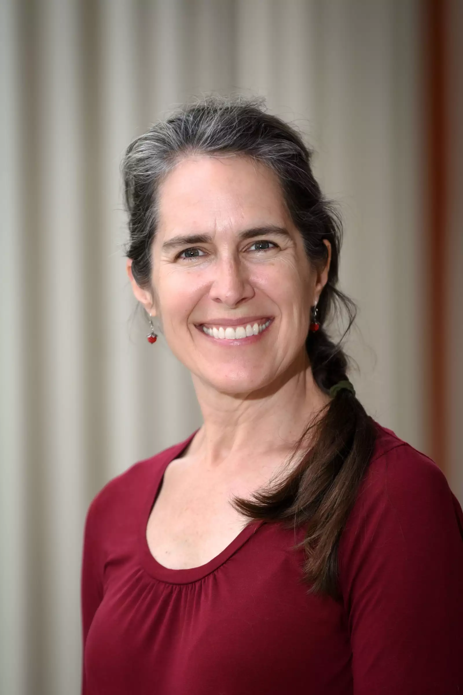 A woman with long dark hair, partially gray, smiles at the camera. She is wearing a long-sleeve maroon shirt and small earrings. The background is blurred with soft vertical lines.