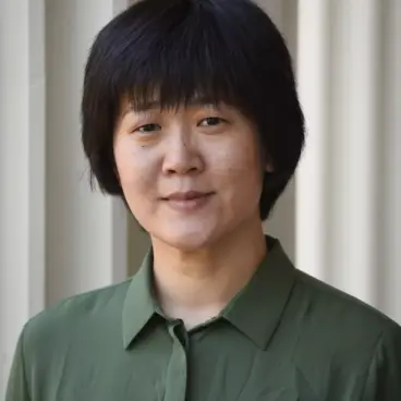 A person with short black hair is wearing a green collared shirt and standing in front of a light-colored wall.