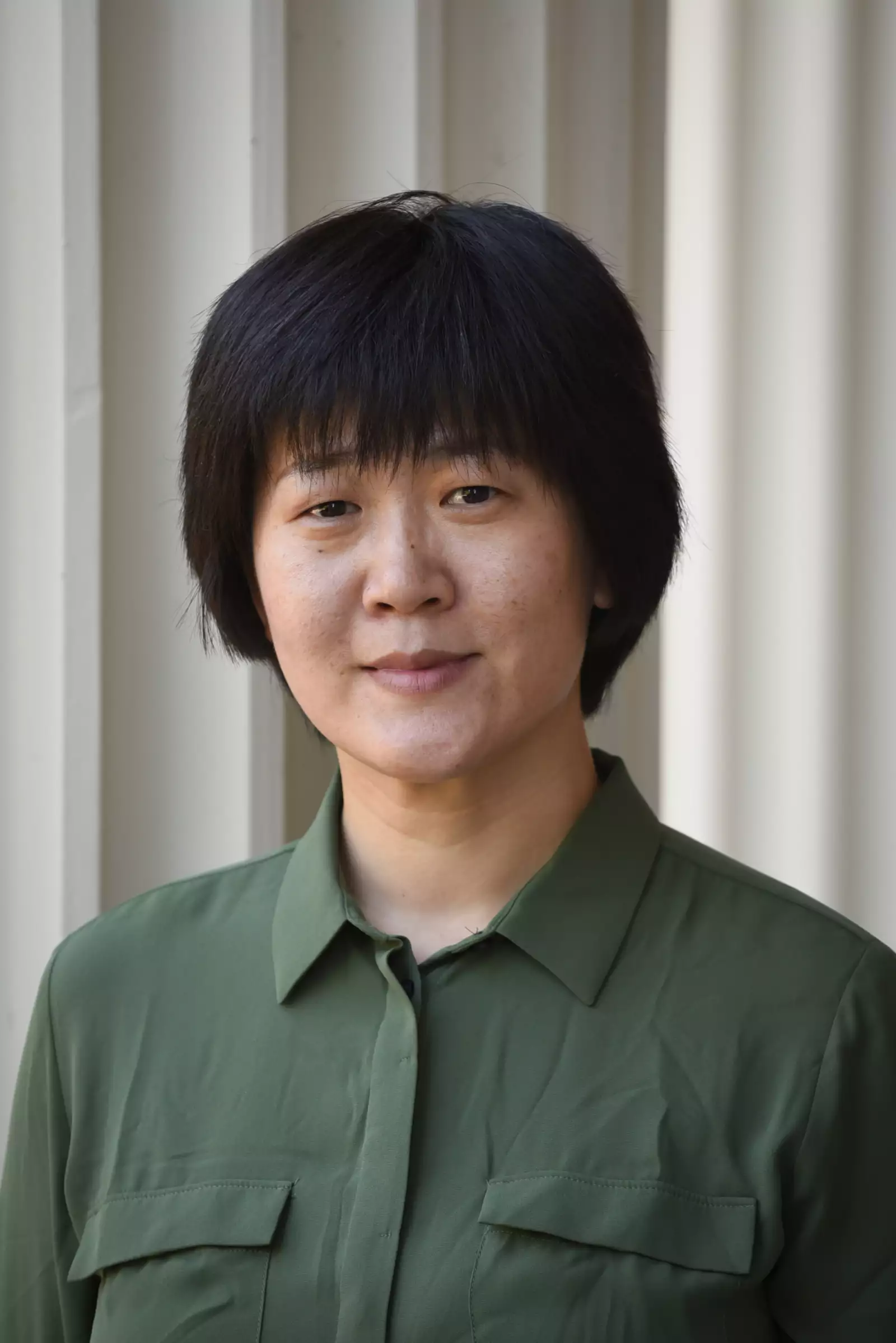 A person with short black hair is wearing a green collared shirt and standing in front of a light-colored wall.