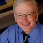 A smiling person with glasses and gray hair is wearing a blue shirt and a black tie with white lettering. The background appears to be an office setting with blurred objects.
