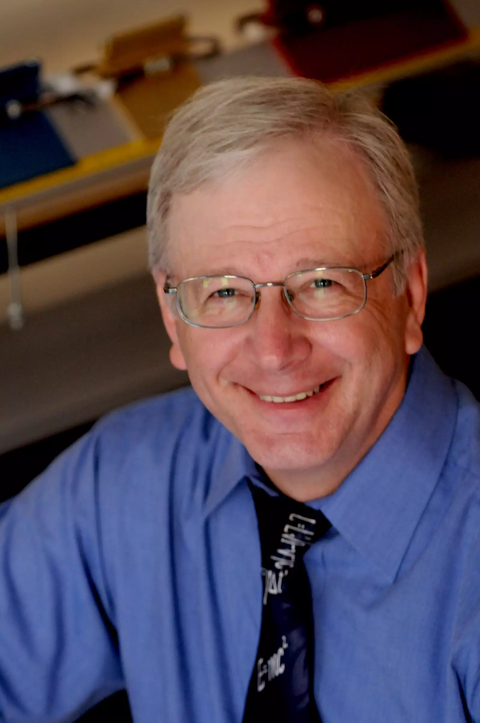 A smiling person with glasses and gray hair is wearing a blue shirt and a black tie with white lettering. The background appears to be an office setting with blurred objects.