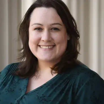 A person with shoulder-length dark hair is smiling at the camera. They are wearing a dark green, patterned top and standing against a light-colored, vertical paneled backdrop.