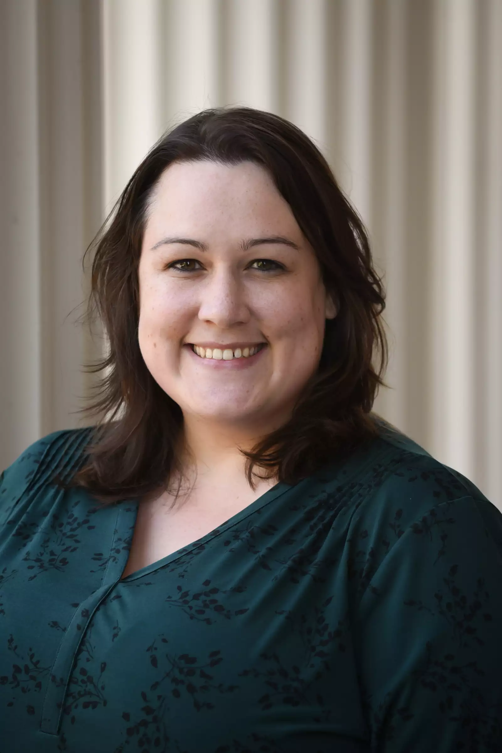 A person with shoulder-length dark hair is smiling at the camera. They are wearing a dark green, patterned top and standing against a light-colored, vertical paneled backdrop.