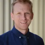 A smiling person with short, light-colored hair, wearing a dark blue button-up shirt and a tie with small polka dots. The background is softly blurred, showing vertical lines.
