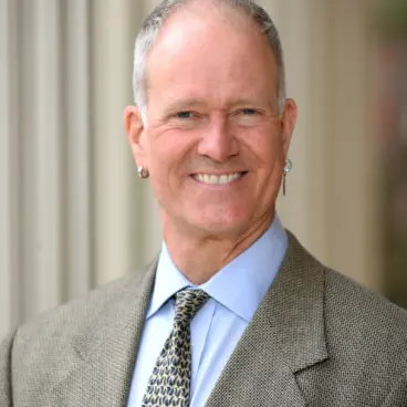 A smiling man in a suit and tie stands in front of a columned building. He has short gray hair and is wearing earrings. The background is softly blurred.
