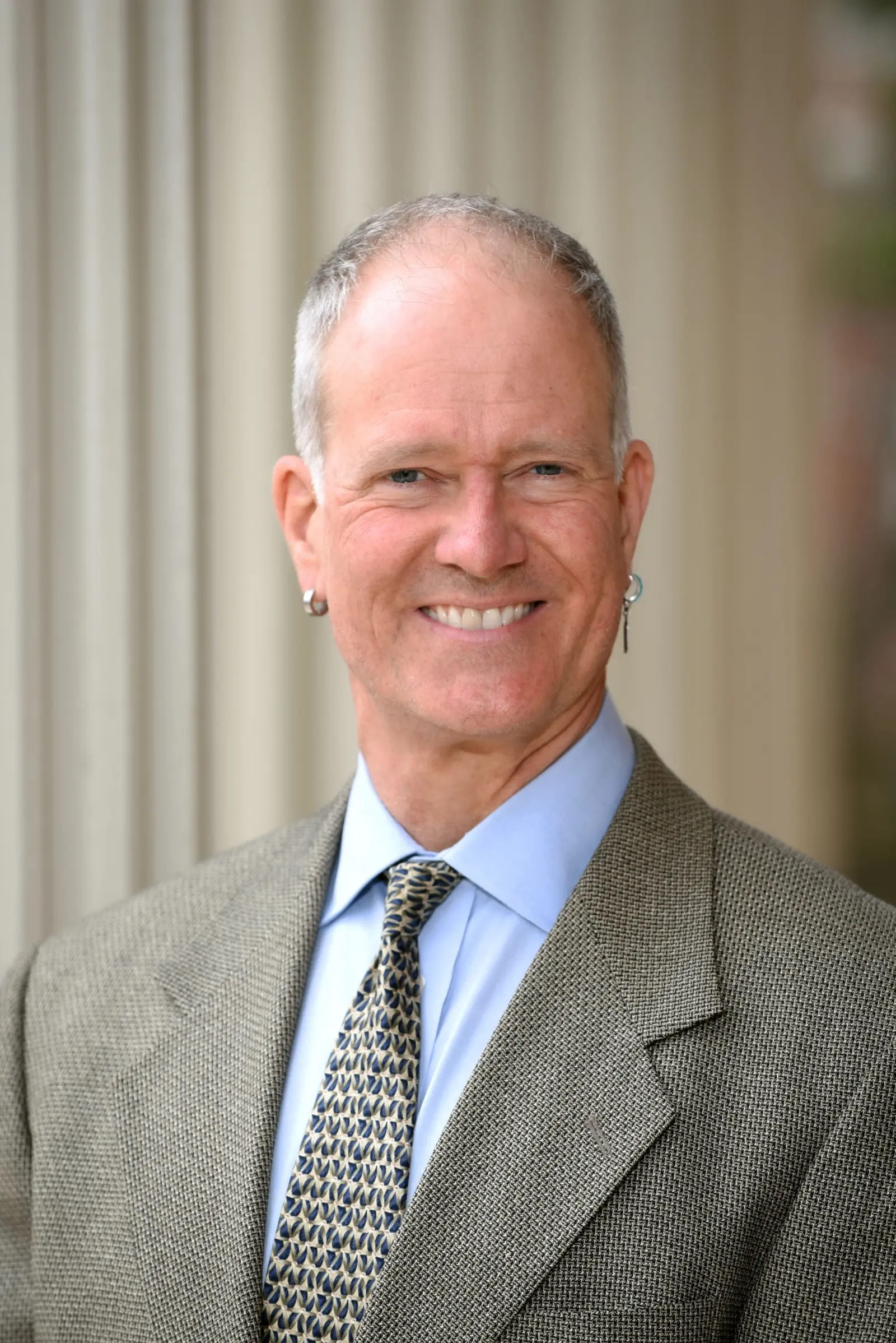 A smiling man in a suit and tie stands in front of a columned building. He has short gray hair and is wearing earrings. The background is softly blurred.