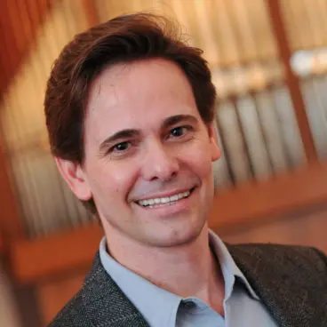 A man in a suit jacket and light blue shirt smiles at the camera. The background features a set of organ pipes.