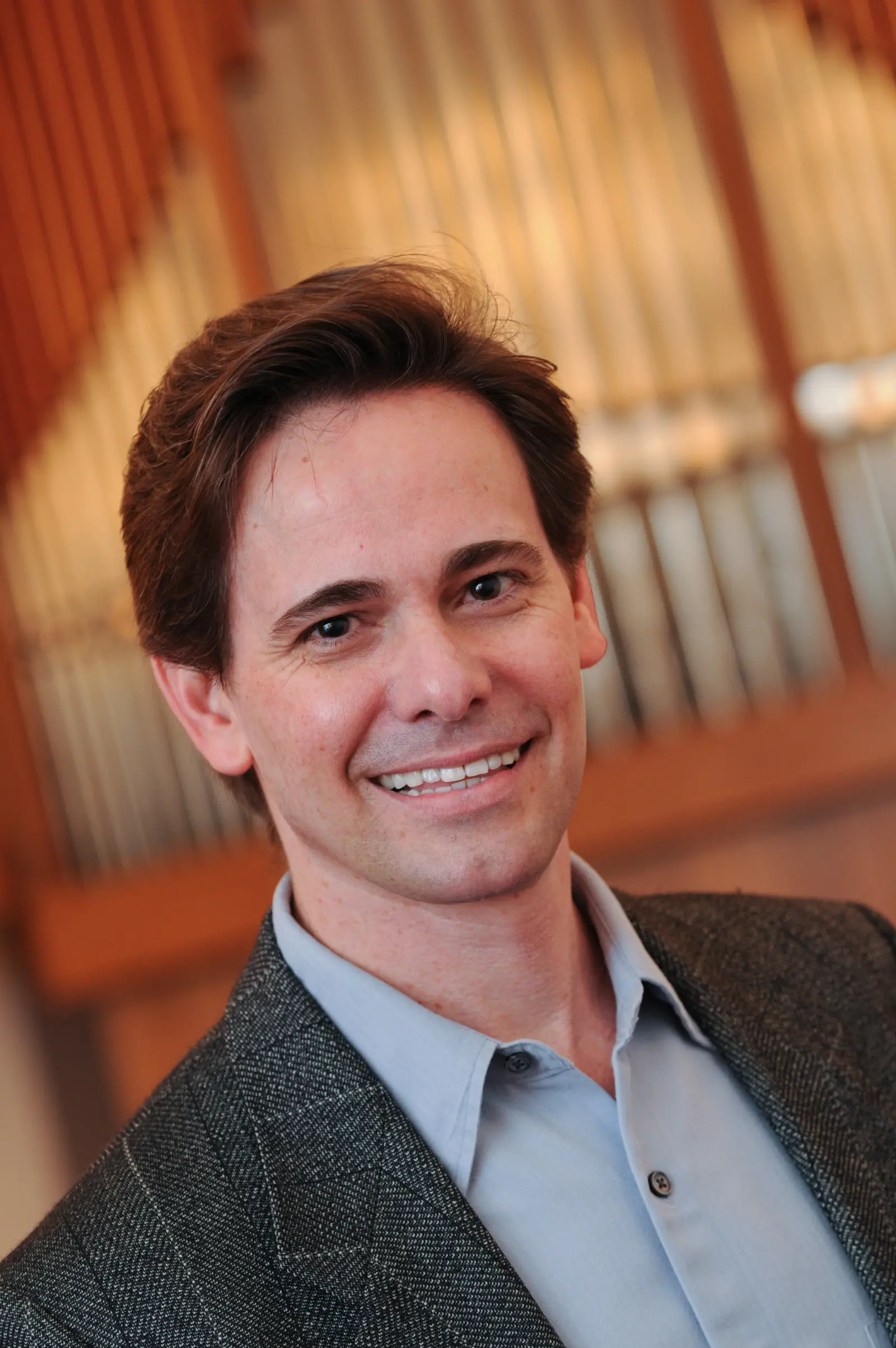 A man in a suit jacket and light blue shirt smiles at the camera. The background features a set of organ pipes.