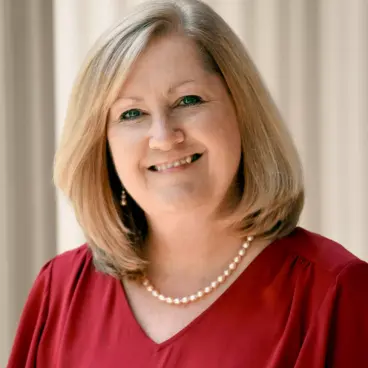 A woman with shoulder-length blonde hair smiling, wearing a red blouse and a pearl necklace. The background is softly blurred, highlighting her friendly expression.