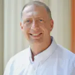 A man wearing a white button-up shirt smiles at the camera. The background features vertical, light-colored lines.