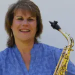 A woman wearing a blue floral blouse is smiling while holding a saxophone. The background is a plain light color.
