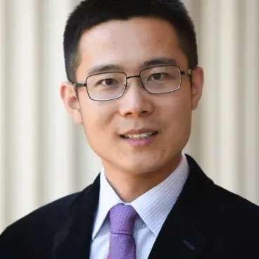 A man with short dark hair and glasses is wearing a black suit, white shirt, and purple tie. He is smiling and facing the camera. The background is softly blurred with vertical white columns.