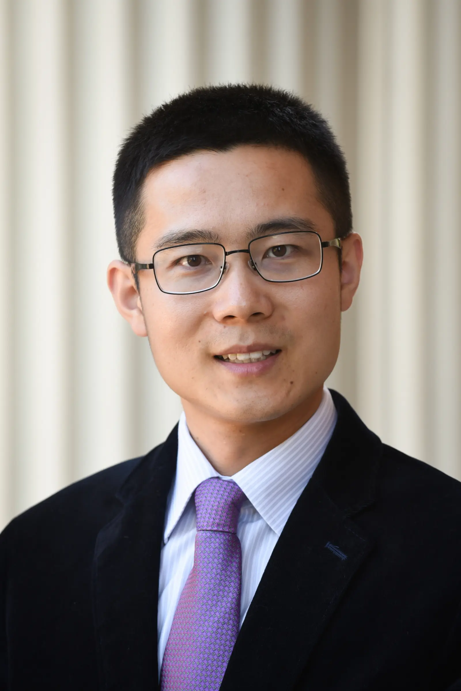A man with short dark hair and glasses is wearing a black suit, white shirt, and purple tie. He is smiling and facing the camera. The background is softly blurred with vertical white columns.