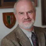 A smiling man with a gray beard and wearing a tweed jacket and patterned tie stands indoors. Behind him, there is a wooden shield with a coat of arms and a framed certificate on the wall.