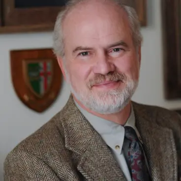 A smiling man with a gray beard and wearing a tweed jacket and patterned tie stands indoors. Behind him, there is a wooden shield with a coat of arms and a framed certificate on the wall.