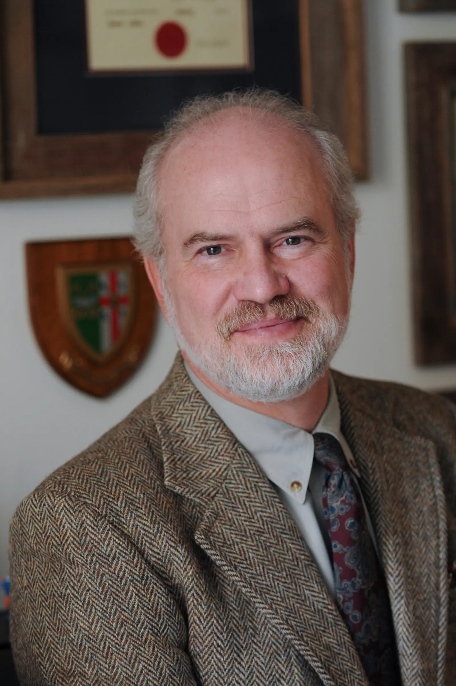 A smiling man with a gray beard and wearing a tweed jacket and patterned tie stands indoors. Behind him, there is a wooden shield with a coat of arms and a framed certificate on the wall.