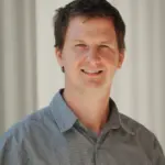 A person with short brown hair smiles while wearing a gray checkered shirt. The background is a neutral, light-colored wall with vertical lines.