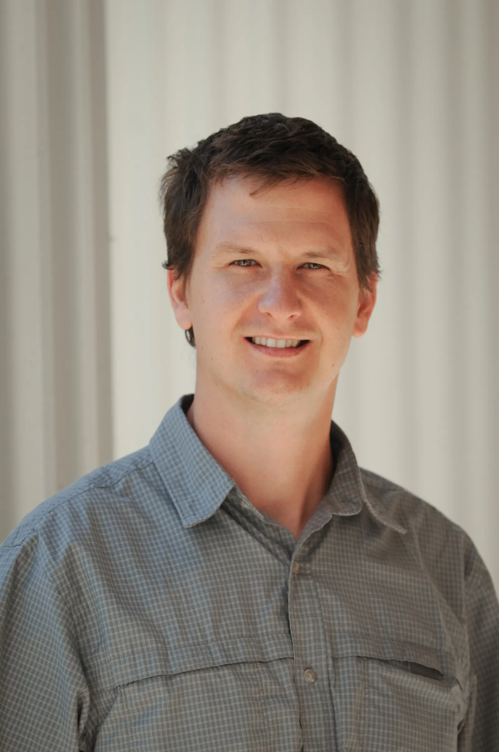 A person with short brown hair smiles while wearing a gray checkered shirt. The background is a neutral, light-colored wall with vertical lines.