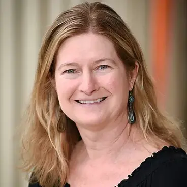 A woman with shoulder-length blonde hair smiles at the camera. She is wearing a black top and dangling earrings. The background is softly blurred, emphasizing her friendly expression.