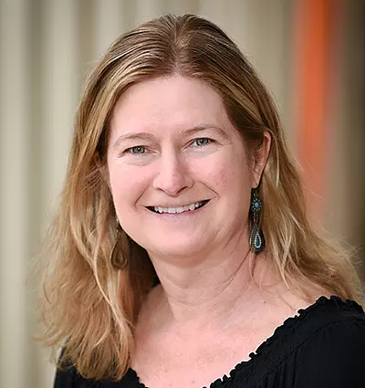 A woman with shoulder-length blonde hair smiles at the camera. She is wearing a black top and dangling earrings. The background is softly blurred, emphasizing her friendly expression.