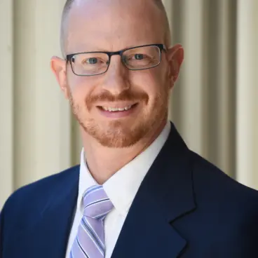 A man with a shaved head and glasses is smiling. He is wearing a dark suit, a light striped tie, and a light-colored shirt. The background is composed of vertical, light-colored blinds.