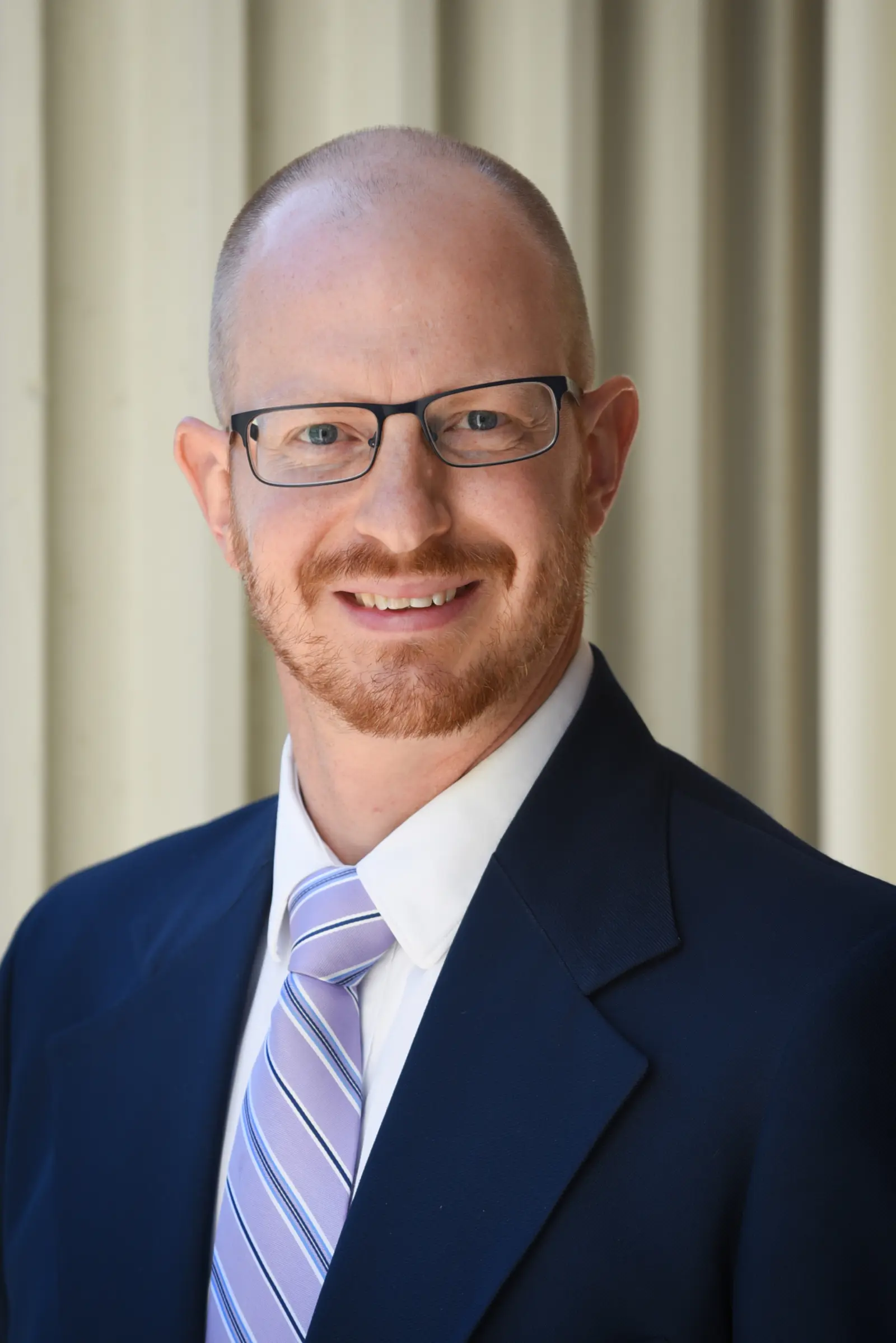 A man with a shaved head and glasses is smiling. He is wearing a dark suit, a light striped tie, and a light-colored shirt. The background is composed of vertical, light-colored blinds.