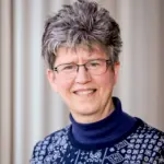 A person with short gray hair and glasses smiles at the camera. They are wearing a navy blue turtleneck with a patterned sweater. The background is softly blurred.