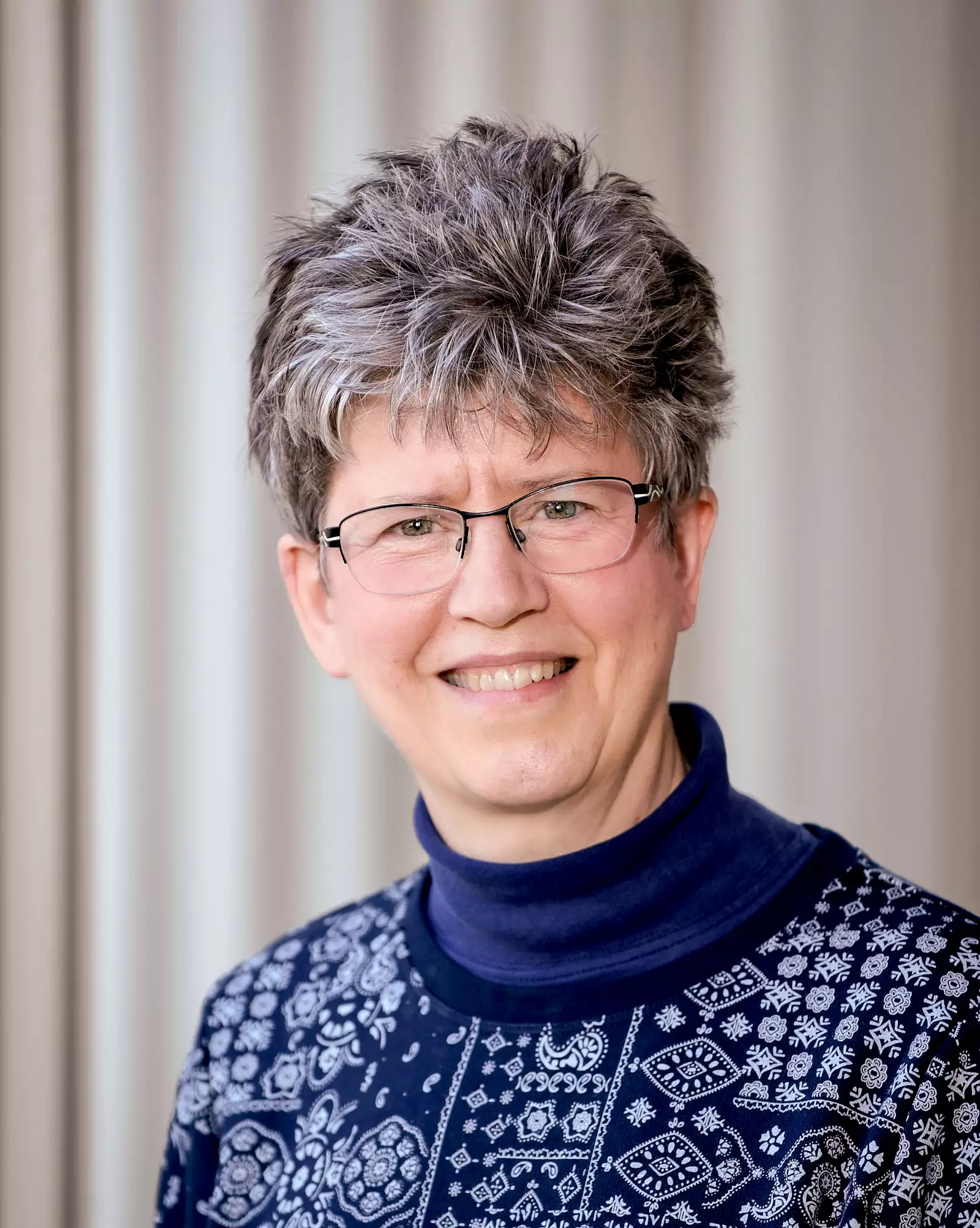 A person with short gray hair and glasses smiles at the camera. They are wearing a navy blue turtleneck with a patterned sweater. The background is softly blurred.