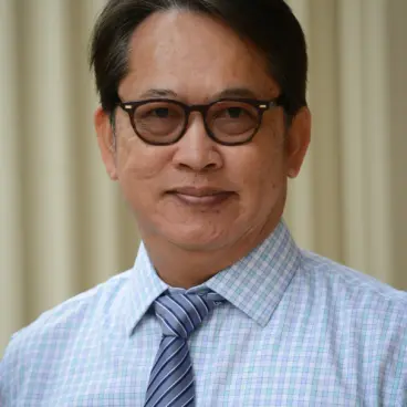 A person with short hair and glasses is wearing a blue and white checkered shirt with a striped tie. The background is blurred beige vertical lines.