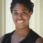 A person with dark hair styled in twists is smiling at the camera. They are wearing long, dangling earrings and a sleeveless black top. The background is softly lit and blurred.