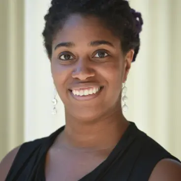 A person with dark hair styled in twists is smiling at the camera. They are wearing long, dangling earrings and a sleeveless black top. The background is softly lit and blurred.
