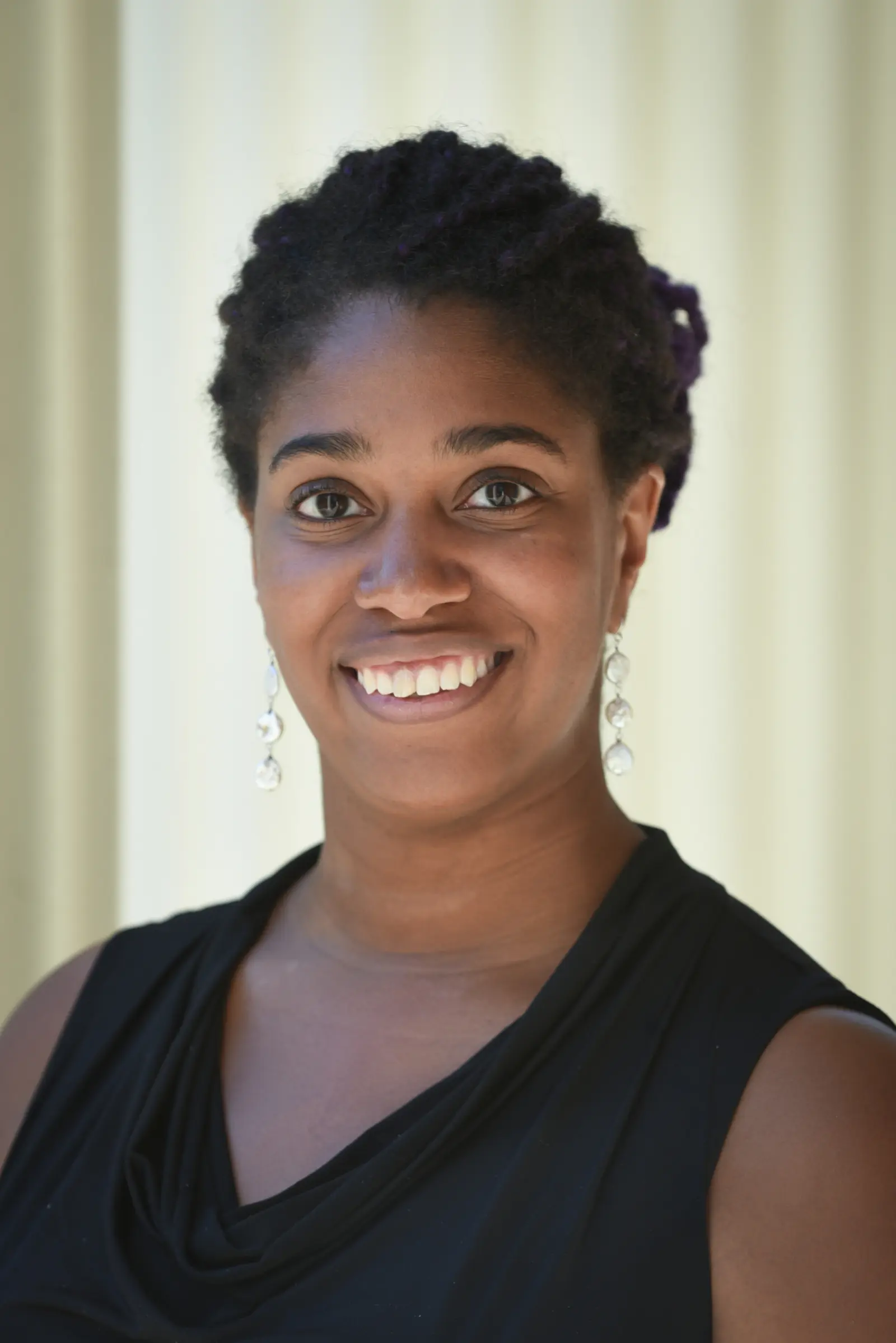 A person with dark hair styled in twists is smiling at the camera. They are wearing long, dangling earrings and a sleeveless black top. The background is softly lit and blurred.