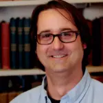 A man with glasses, wearing a light blue shirt, smiles in front of a bookshelf filled with a variety of books.