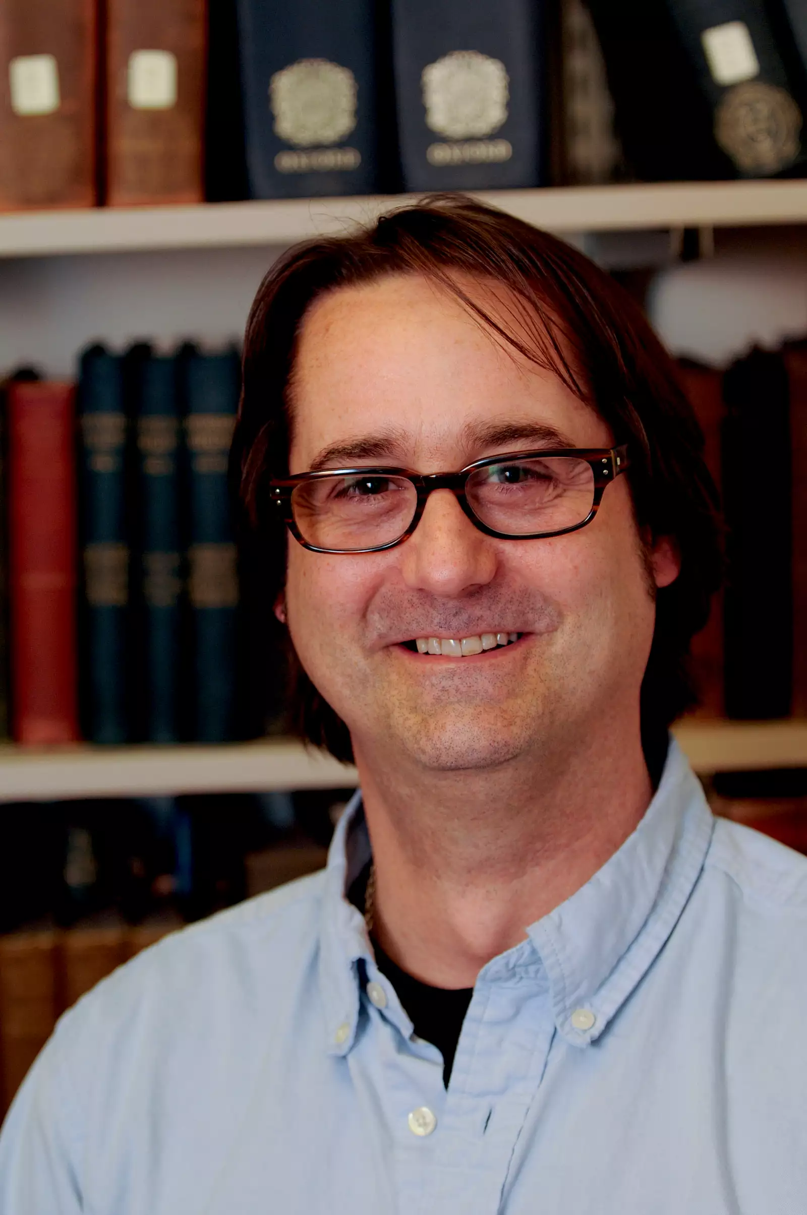 A man with glasses, wearing a light blue shirt, smiles in front of a bookshelf filled with a variety of books.