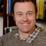 A smiling man with short dark hair, wearing a plaid shirt under a brown sweater, is sitting in front of a filled bookshelf.