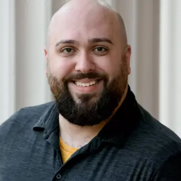 A bald man with a beard smiles at the camera. He is wearing a dark polo shirt and stands against a beige background with vertical lines.