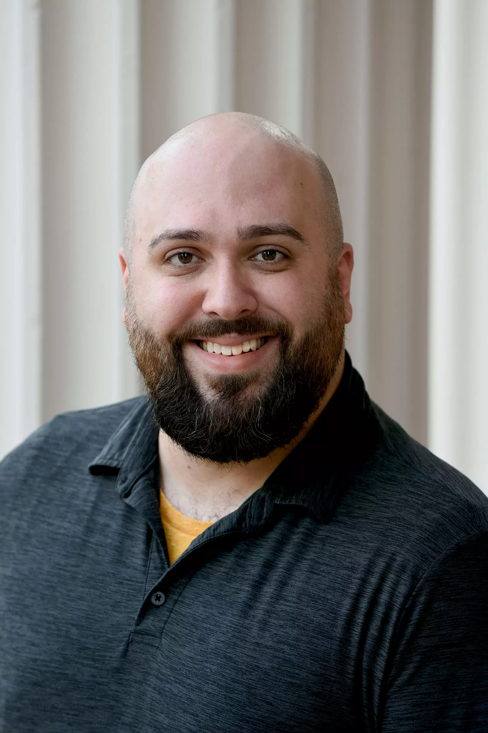 A bald man with a beard smiles at the camera. He is wearing a dark polo shirt and stands against a beige background with vertical lines.