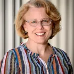 A person with short blonde hair and glasses is smiling. They are wearing a striped, multicolored blouse and standing in front of a light-colored, corrugated background.