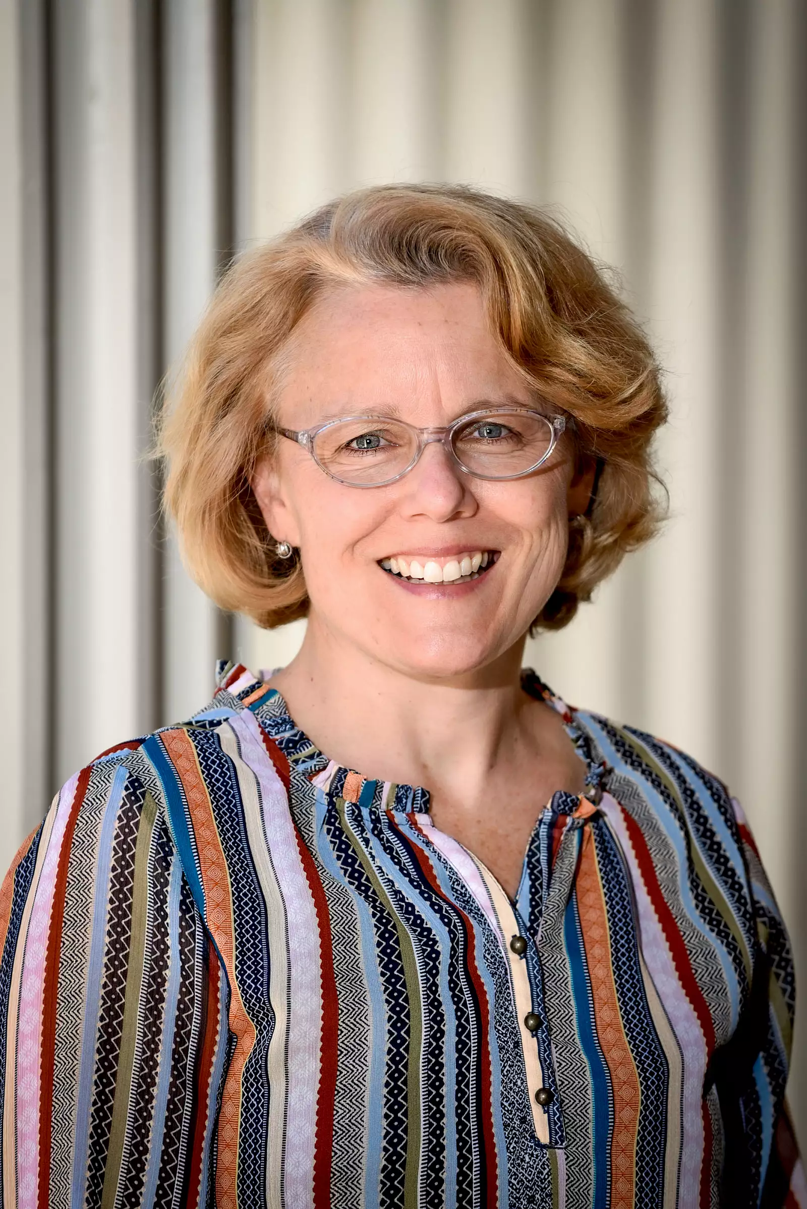 A person with short blonde hair and glasses is smiling. They are wearing a striped, multicolored blouse and standing in front of a light-colored, corrugated background.