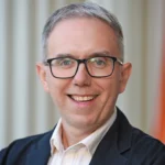 A man with short brown hair and glasses in a dark suit and striped shirt stands in front of a brick building, smiling at the camera.