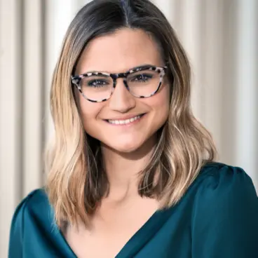 A person with shoulder-length, wavy hair and glasses, smiling while wearing a teal blouse. They are standing in front of a softly lit, neutral-colored background.