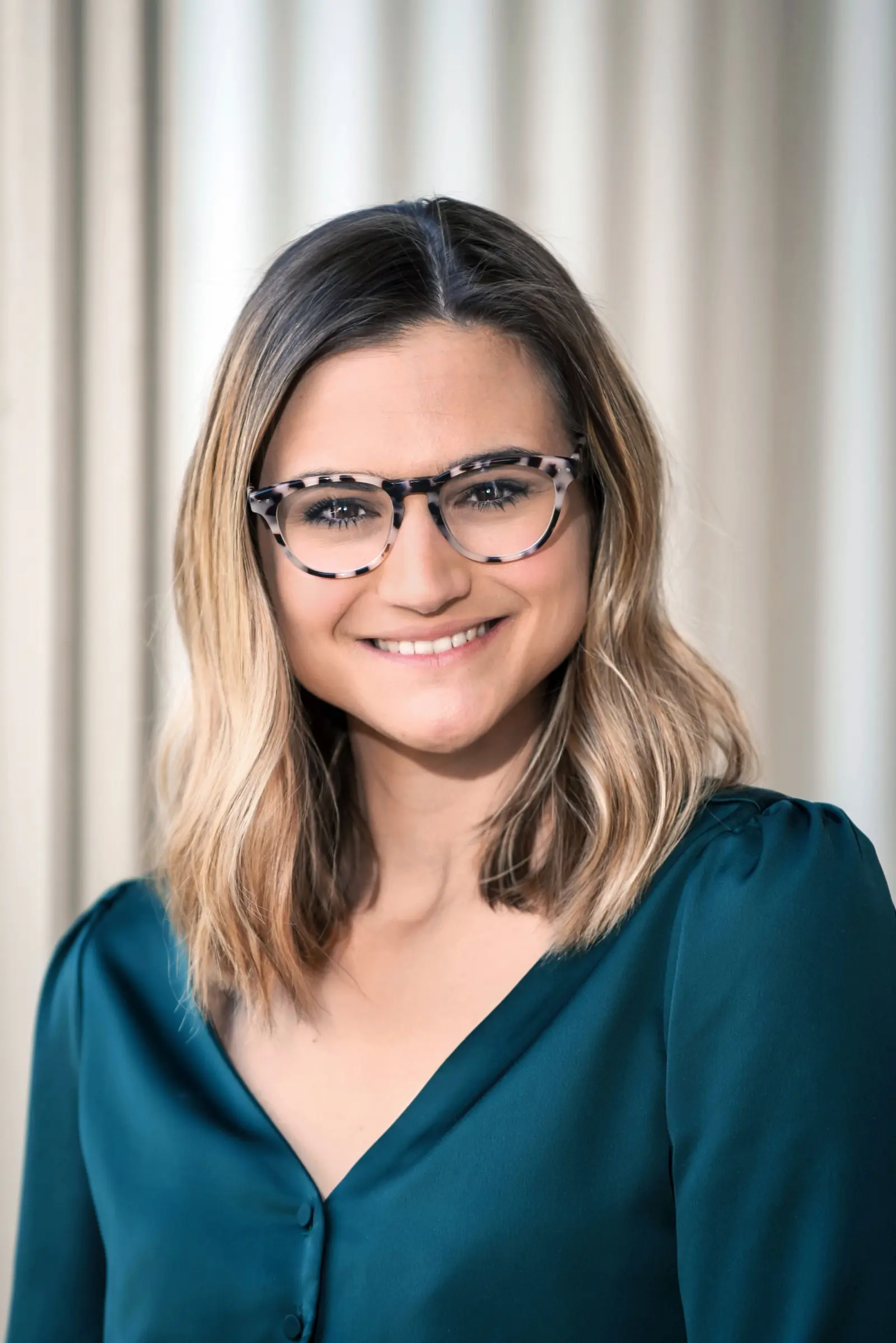 A person with shoulder-length, wavy hair and glasses, smiling while wearing a teal blouse. They are standing in front of a softly lit, neutral-colored background.