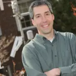 A man with short gray hair and a green button-up shirt stands outdoors with arms crossed, smiling. He is in front of a brick building and some equipment with greenery in the background.