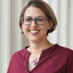 A person with short hair and glasses is smiling, wearing a maroon blouse and a geometric necklace, standing in front of a light-colored, vertically striped wall.