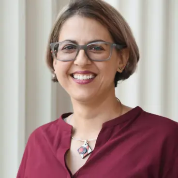 A person with short hair and glasses is smiling, wearing a maroon blouse and a geometric necklace, standing in front of a light-colored, vertically striped wall.